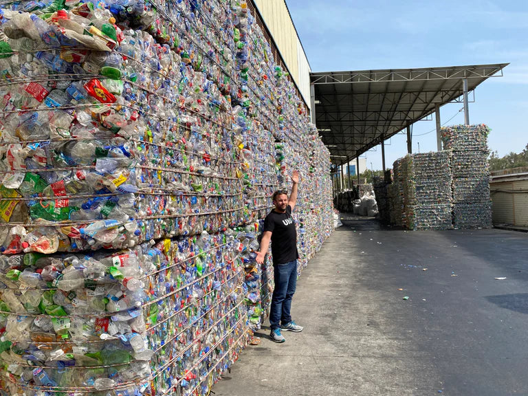 Ben in front of a giant wall of plastic bottles that have been collected from the beaches and are going to be made into ULU bags